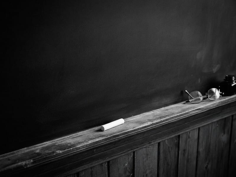 A piece of white chalk, a pair of round spectacles, and an ink bottle rest on the wooden ledge of a blank chalkboard.