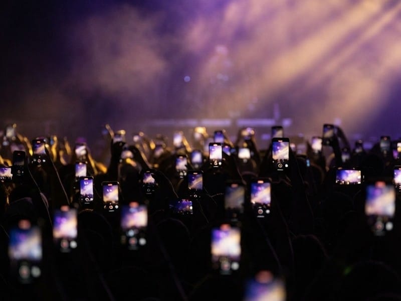 A crowd of people in silhouette holding up illuminated smartphones to record a concert stage under purple and gold stage lights.