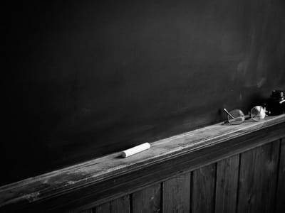 A piece of white chalk, a pair of round spectacles, and an ink bottle rest on the wooden ledge of a blank chalkboard.