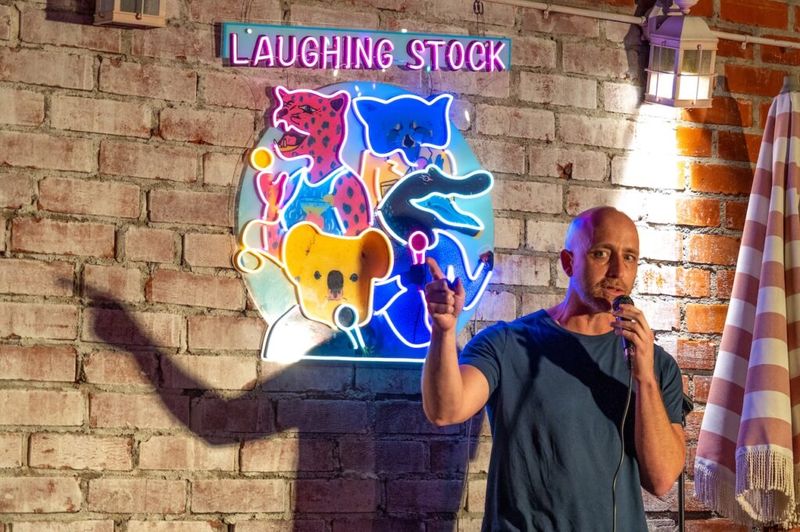 A comedian performs stand-up beside the neon 'Laughing Stock' sign at the Laughs on Larchmont show.